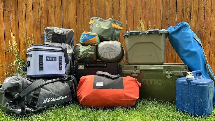 Car camping gear stacked on grass: soft cooler, hard coolers, duffel bags, packed tents, storage bin, folding chair bag and a blue water jug